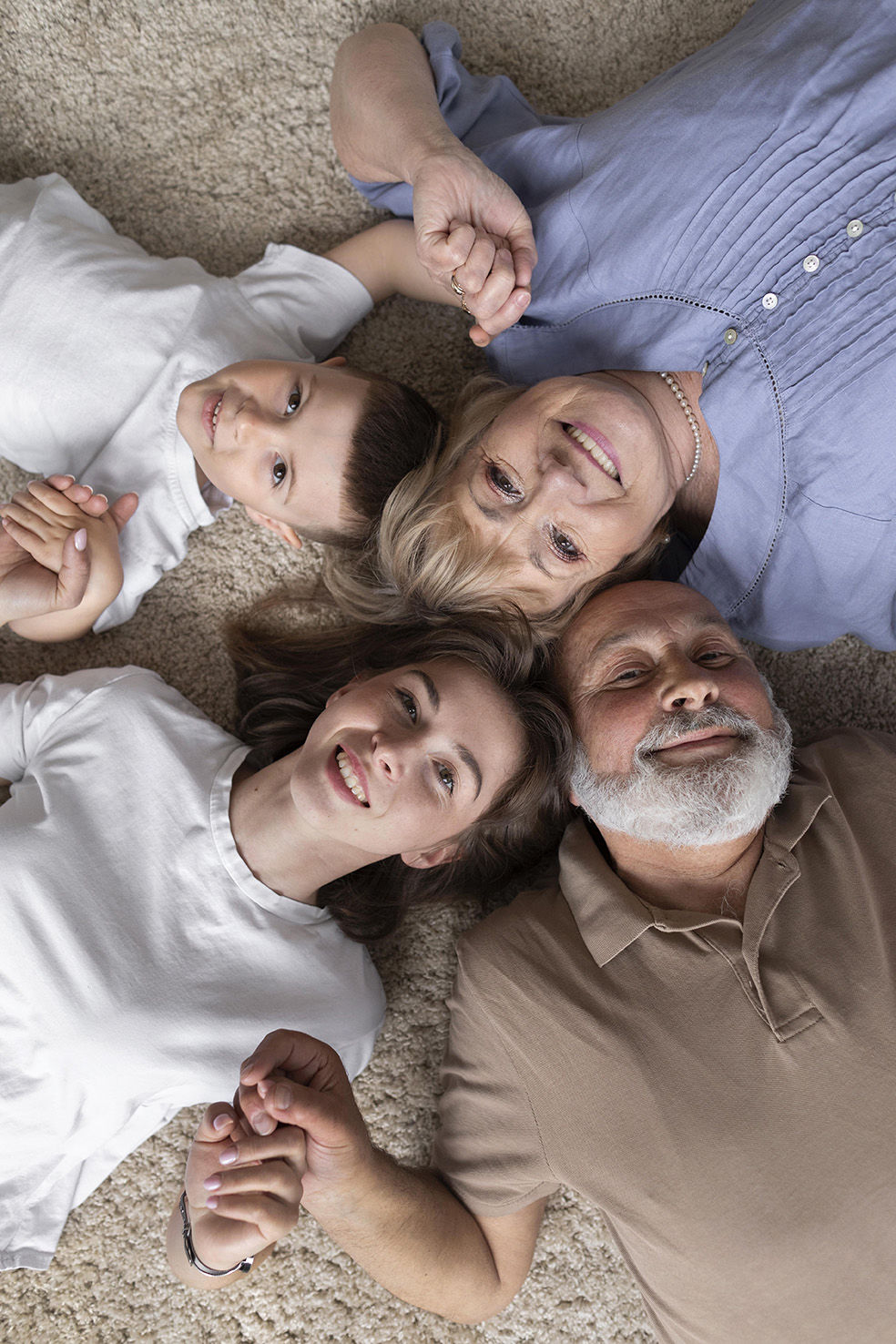 Familia de cuatro generaciones acostada en el suelo sobre una alfombra, formando un círculo y tomados de las manos. Un hombre mayor con barba, una mujer mayor sonriente, una mujer joven con los ojos cerrados y un niño con una expresión juguetona disfrutan de un momento de unión y felicidad.