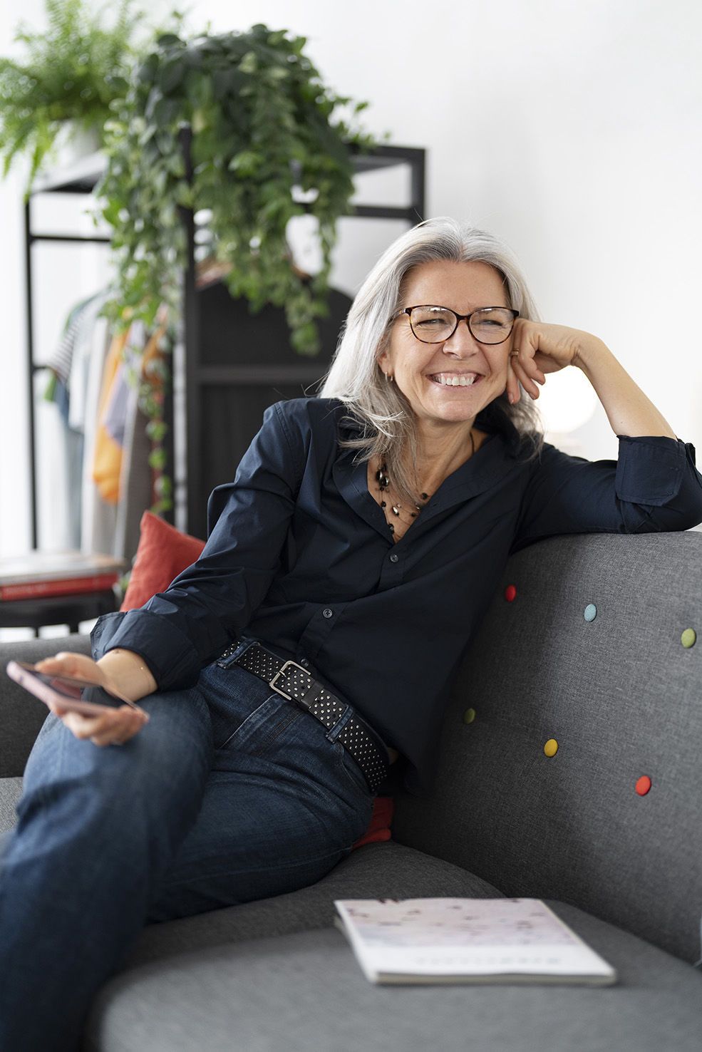 Mujer mayor con cabello canoso y gafas sonríe mientras se recuesta en un sofá gris con botones de colores. Lleva una camisa oscura y jeans con un cinturón decorado, sosteniendo un teléfono en una mano. Sobre la mesa frente a ella hay una revista, en un ambiente moderno y relajado.